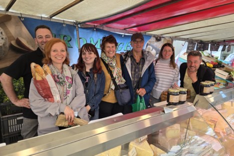 a group of people posing in front of tthe boutique of a cheese vendor at a parisian market